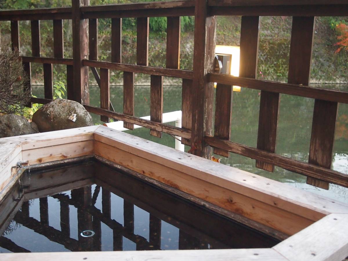 A wooden hot tub fills with water next to a railing overlooking a pond.