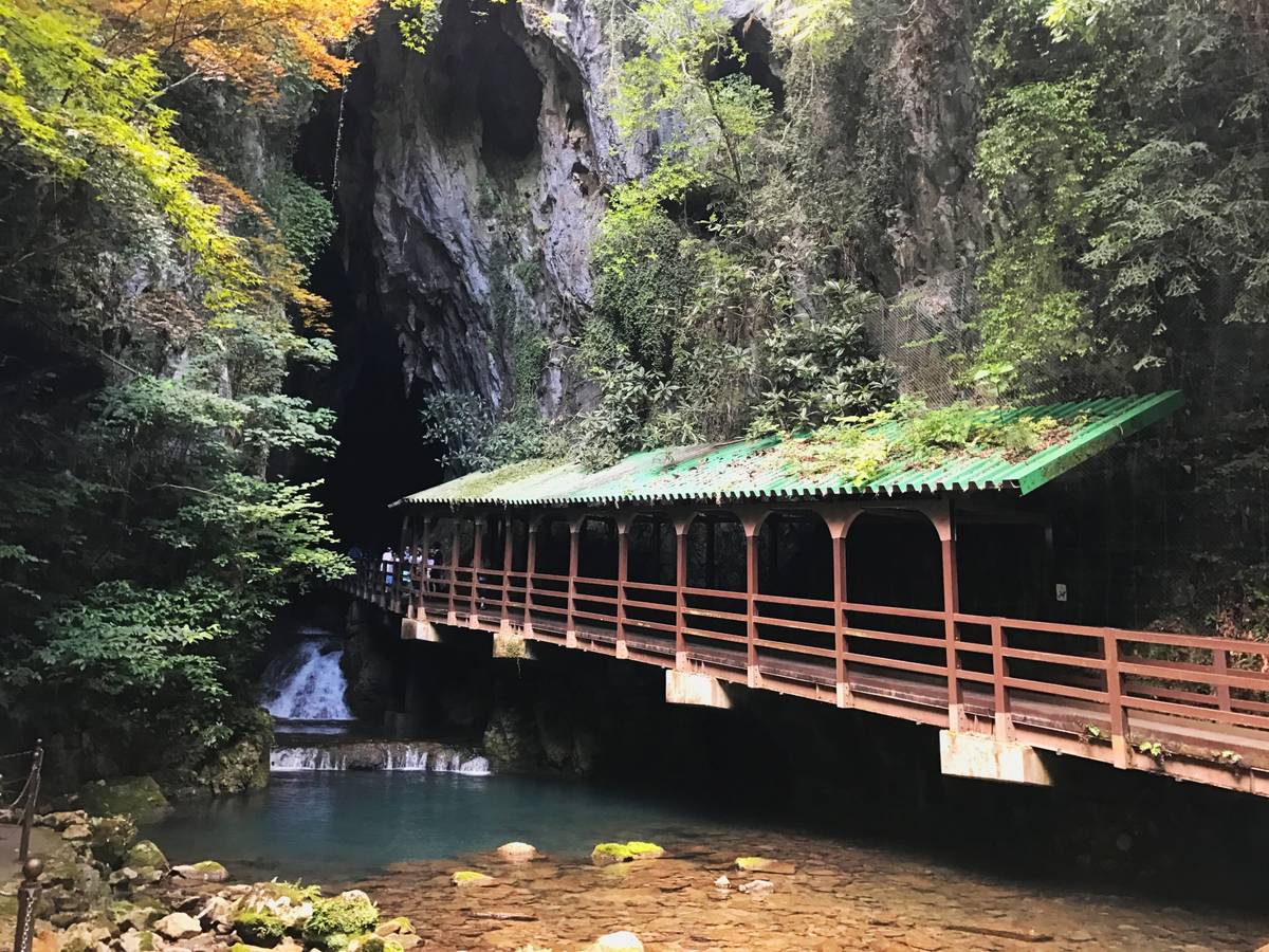 A covered wooden walkway extends over a blue stream towards a dark cave entrance, surrounded.