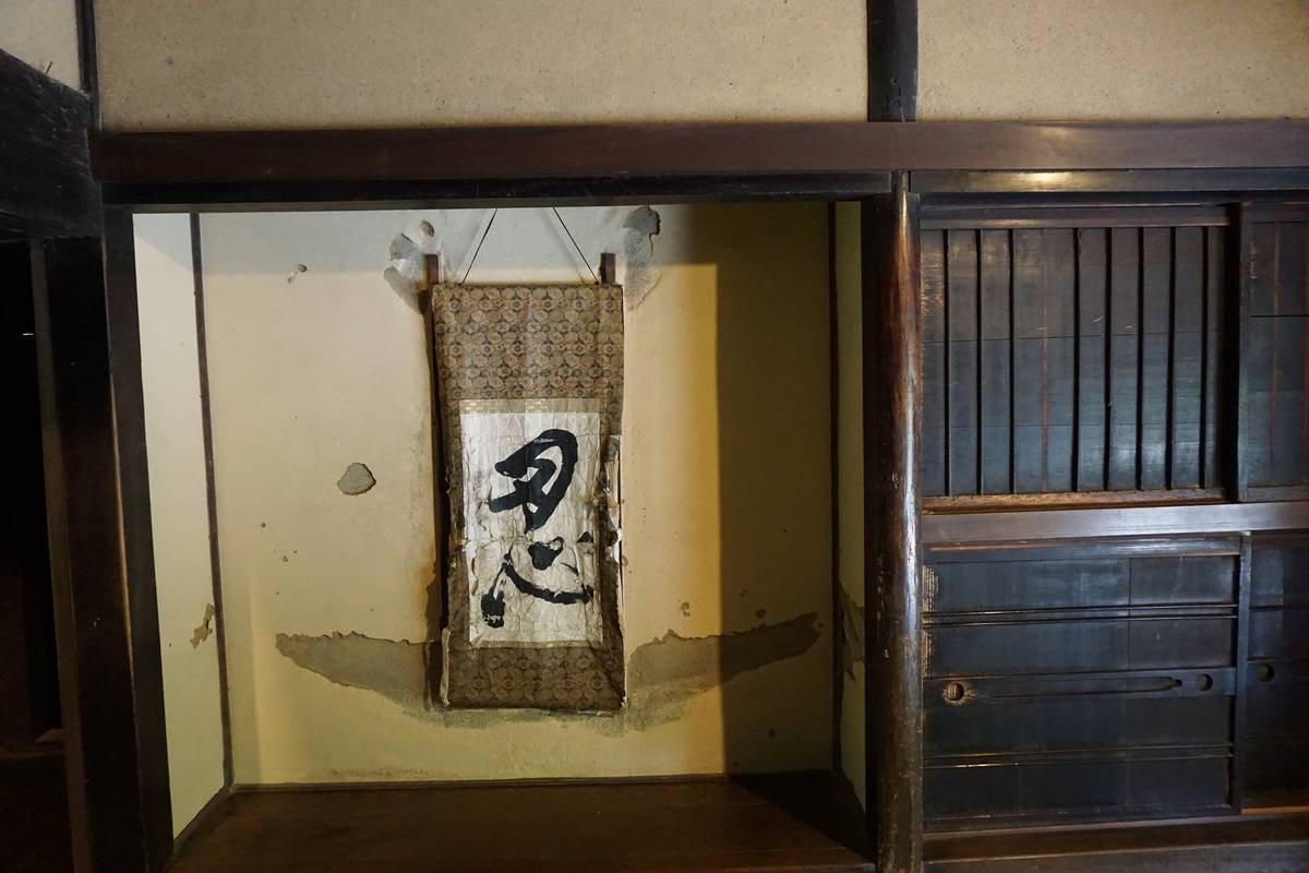 A hanging scroll with a kanji character on a distressed wall, next to a dark wooden sliding door