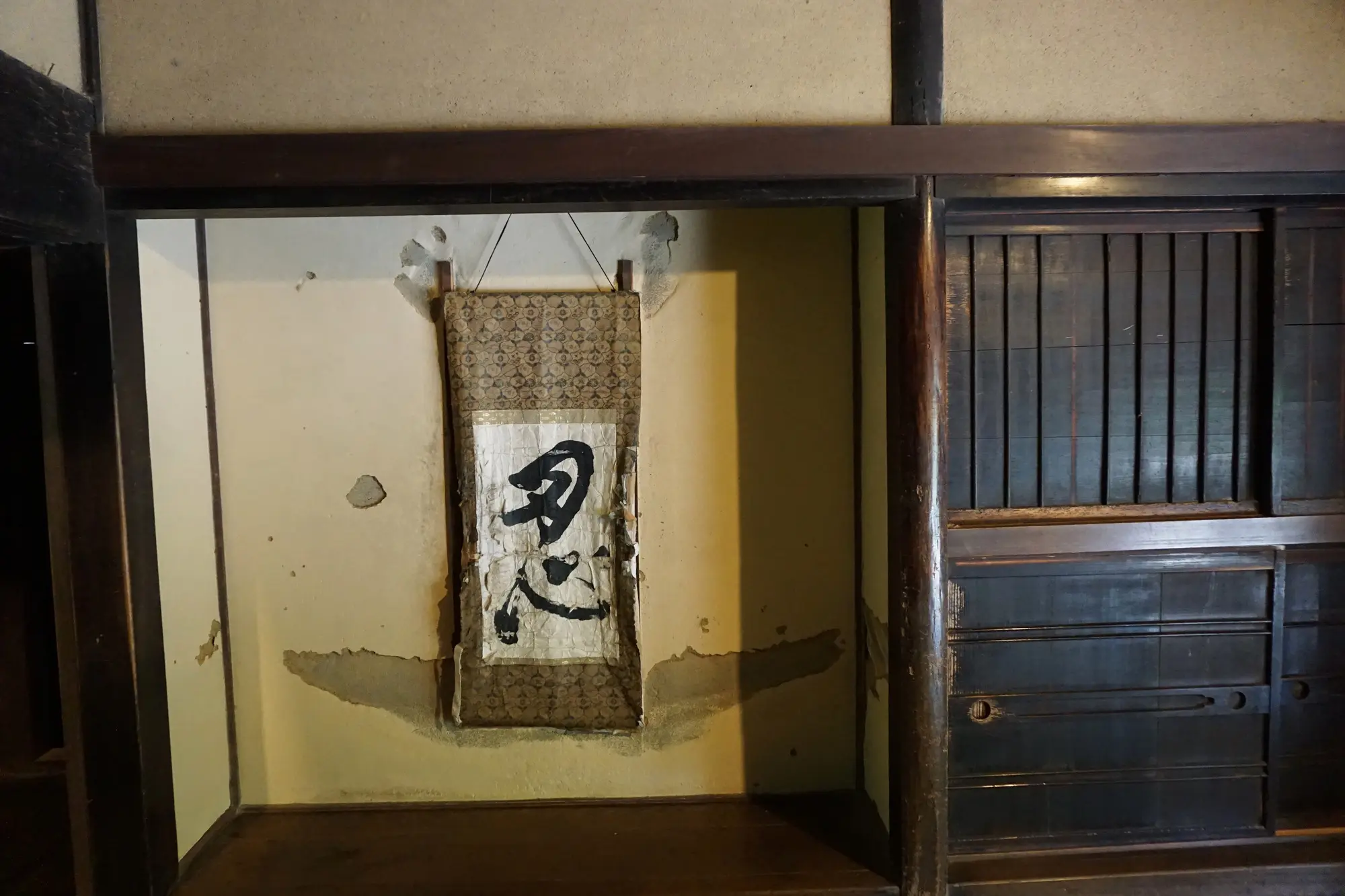 A hanging scroll with a kanji character on a distressed wall, next to a dark wooden sliding door