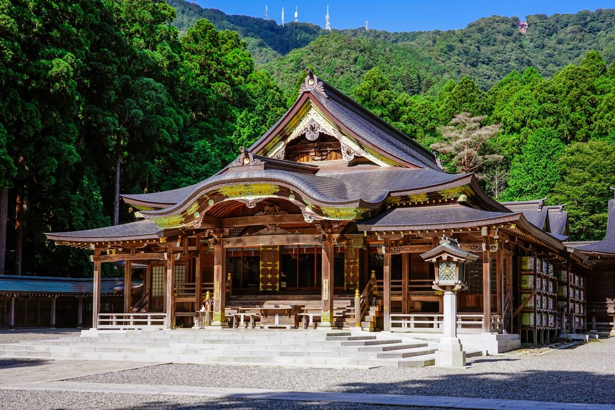 An ornate wooden Japanese temple with curved dark roofs and a stone lantern, set against a backdrop of lush green mountains.
