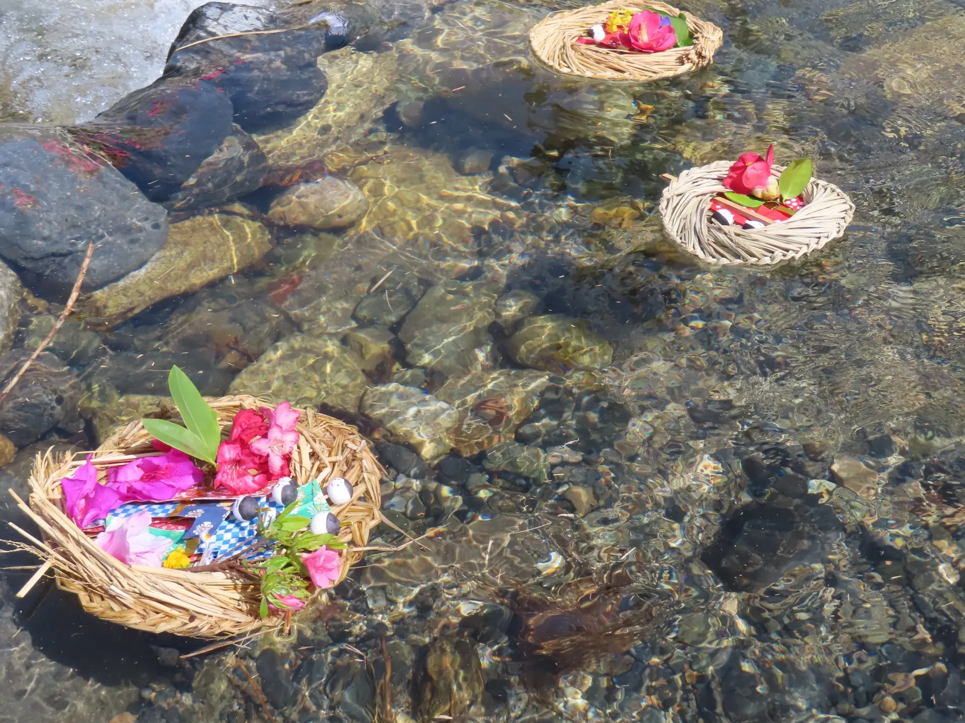 Three woven baskets filled with colorful flowers and offerings floating in clear, shallow water over a rocky riverbed.