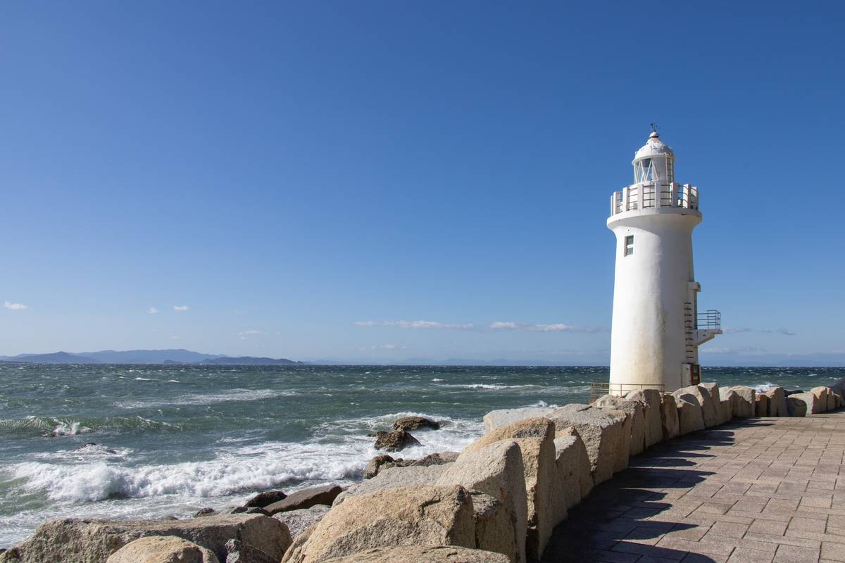 White lighthouse on a rocky shore next to a paved walkway with ocean waves and a clear blue sky.