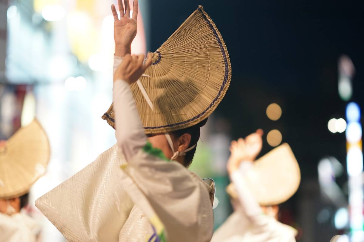 Dancer in white traditional attire and a wide straw hat, seen from behind with an arm raised, at a night festival.