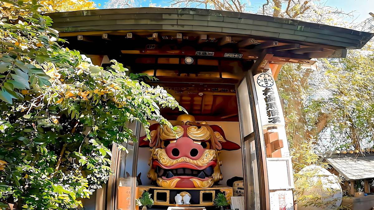 A large, red and gold Japanese lion head mask is displayed in an open wooden shrine.
