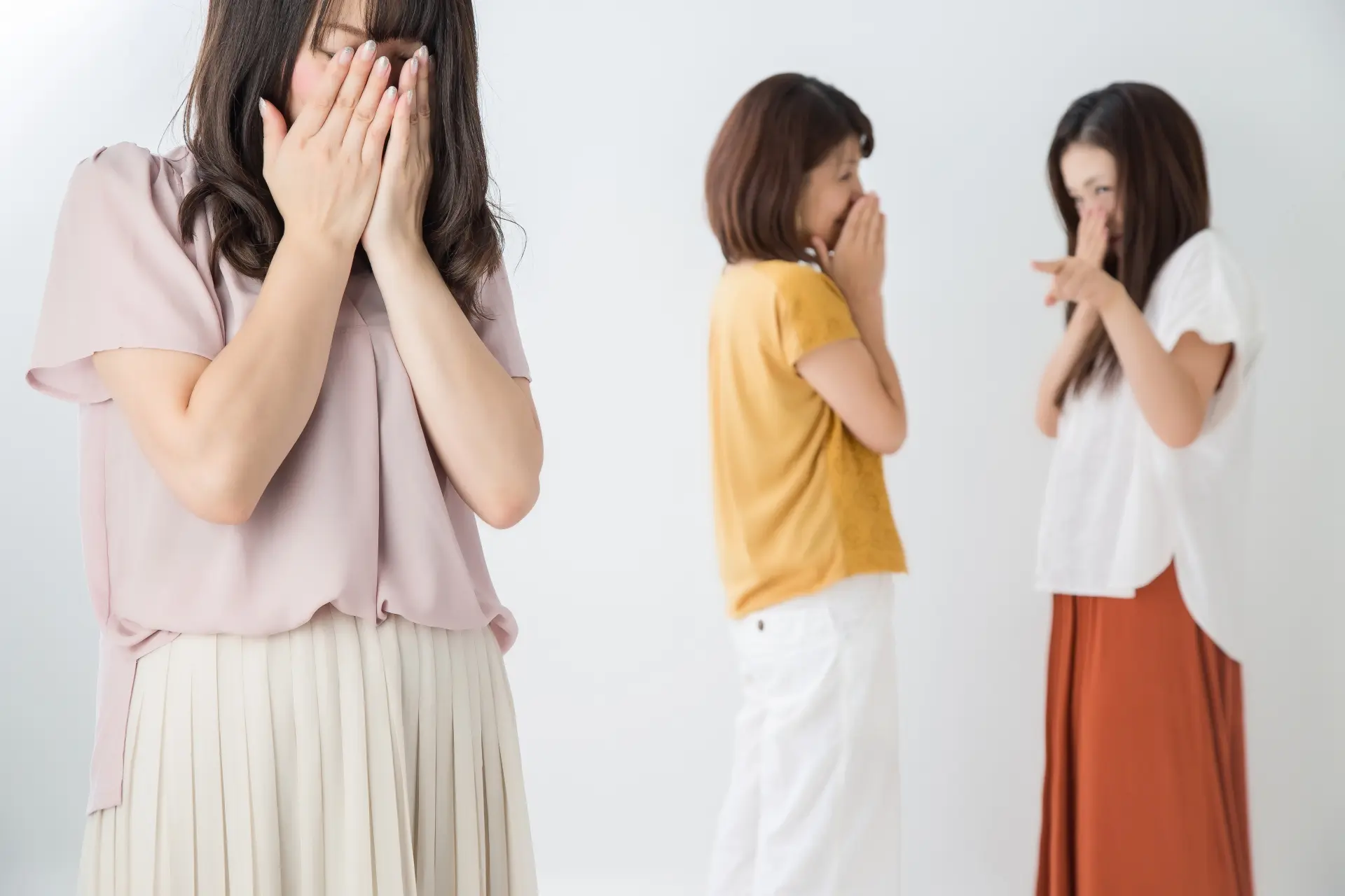 A woman covers her face in distress while two blurred women in the background laugh and point.