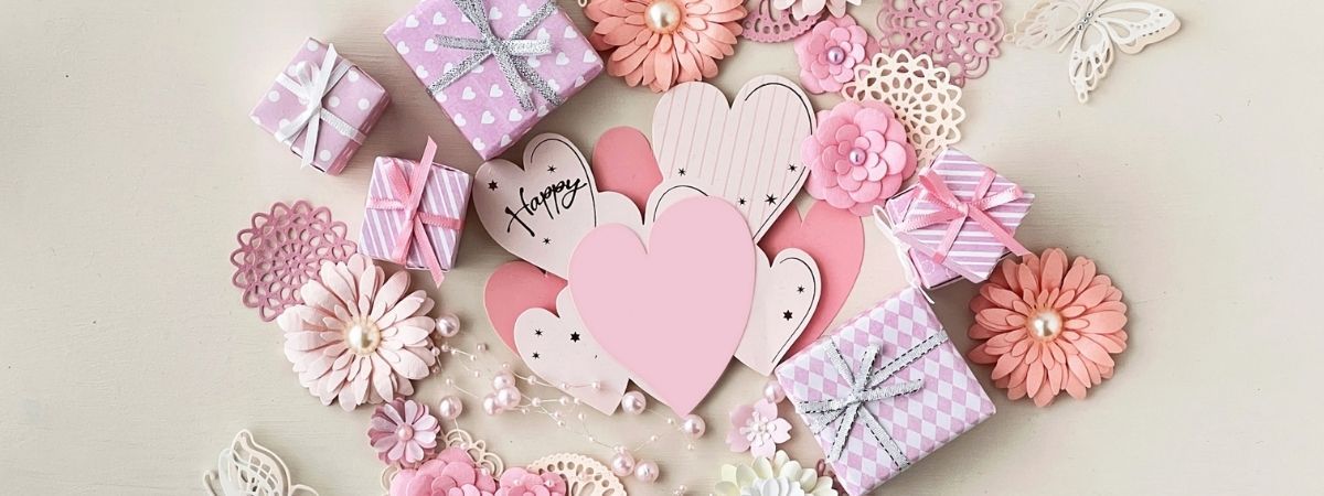 Pastel pink and lavender crafting items including gift boxes, paper hearts, and flowers with scattered pearls.