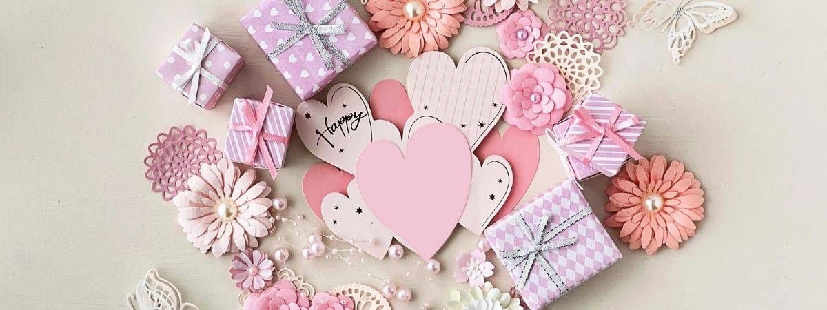 Pastel pink and lavender crafting items including gift boxes, paper hearts, and flowers with scattered pearls.