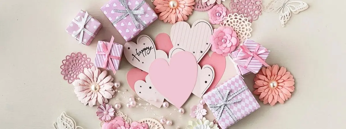 Pastel pink and lavender crafting items including gift boxes, paper hearts, and flowers with scattered pearls.