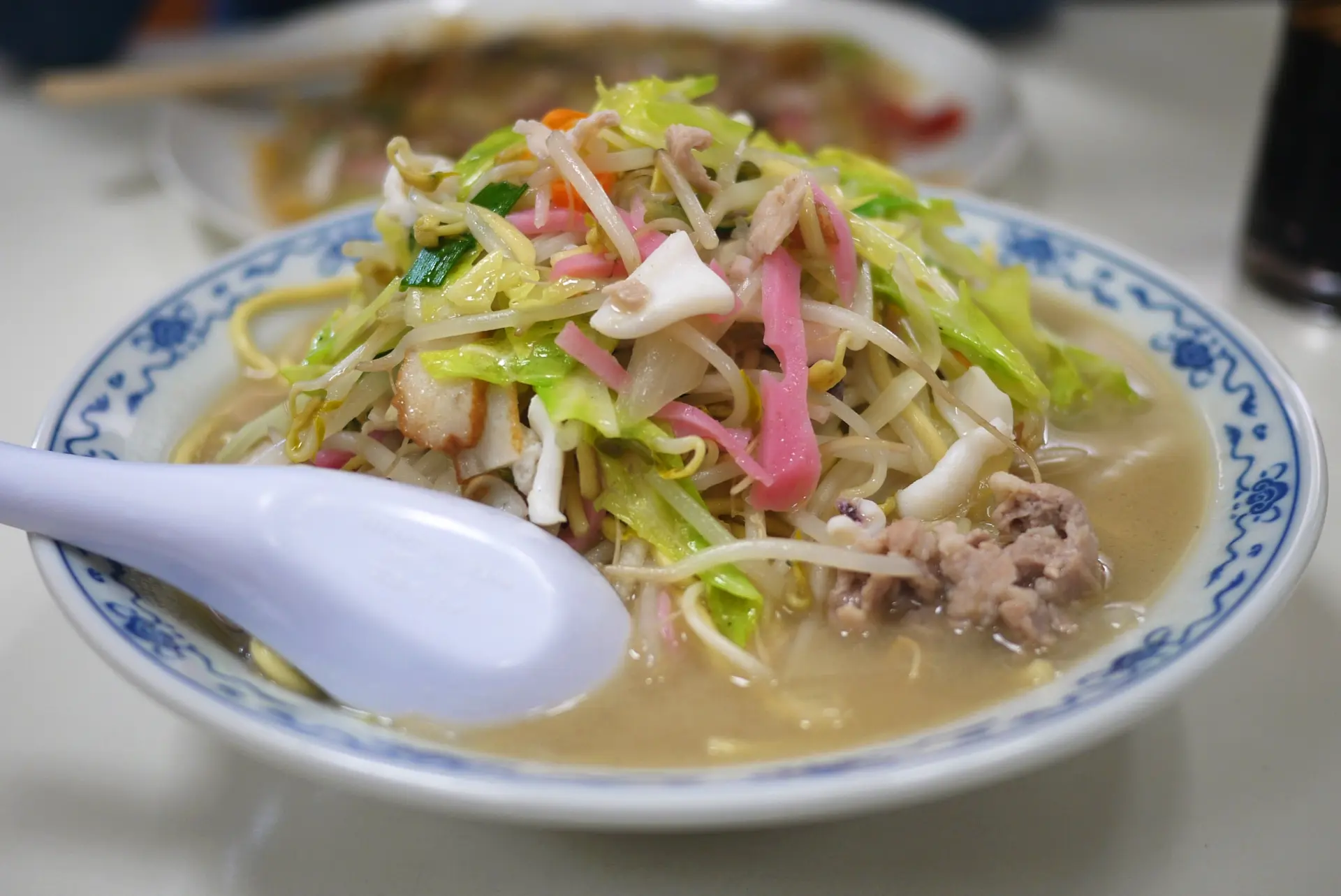 A bowl of Champon noodle soup with cabbage, bean sprouts, meat, squid, and pink fish cake, with a white spoon.