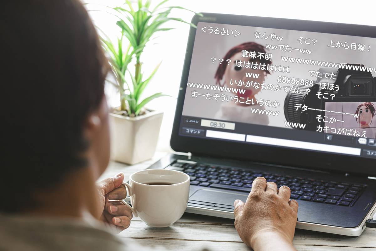 A person holding a coffee cup watches a laptop displaying a live stream with Japanese chat comments.
