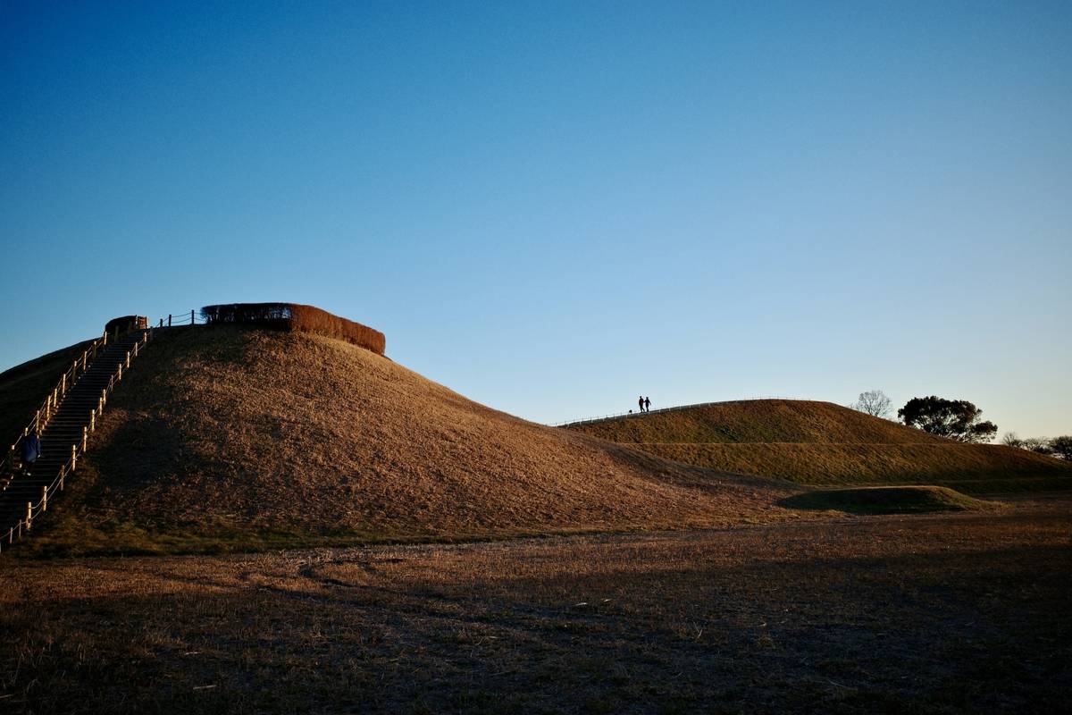 Dry grassy mounds under a clear blue sky, with a staircase leading to a circular structure on the left.