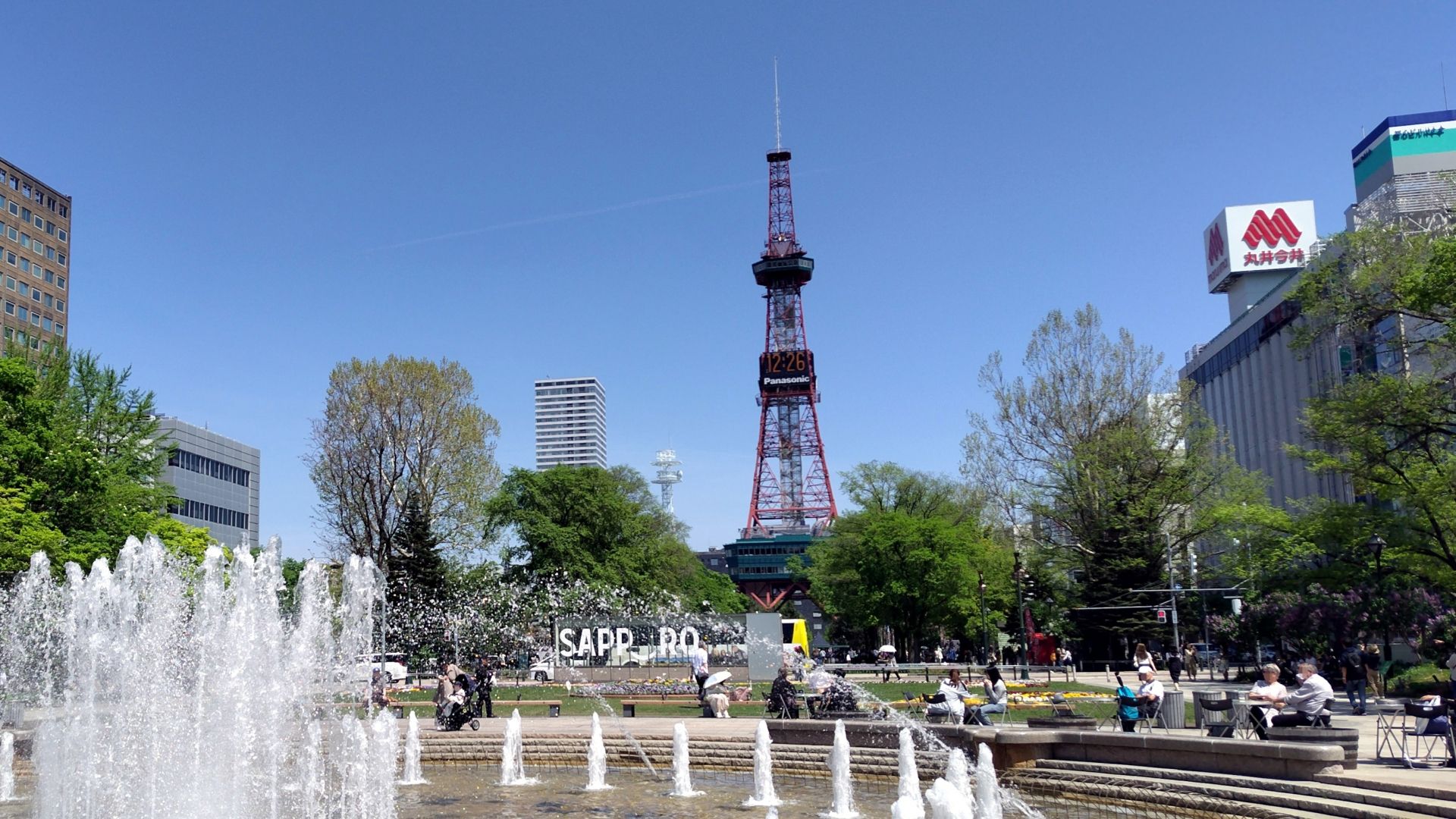 The Sapporo TV Tower stands over a park with fountains and people under a blue sky.