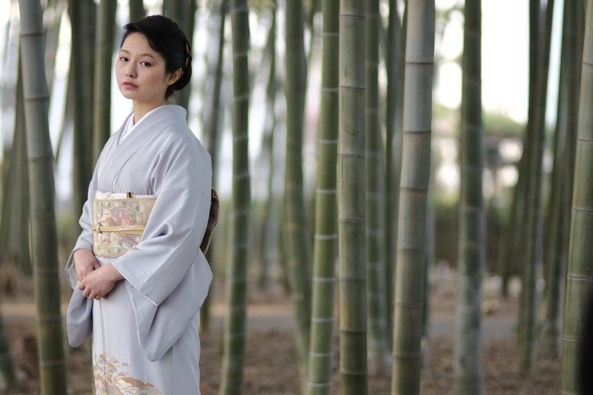 A young woman in a light gray kimono stands in a bamboo forest, looking forward.