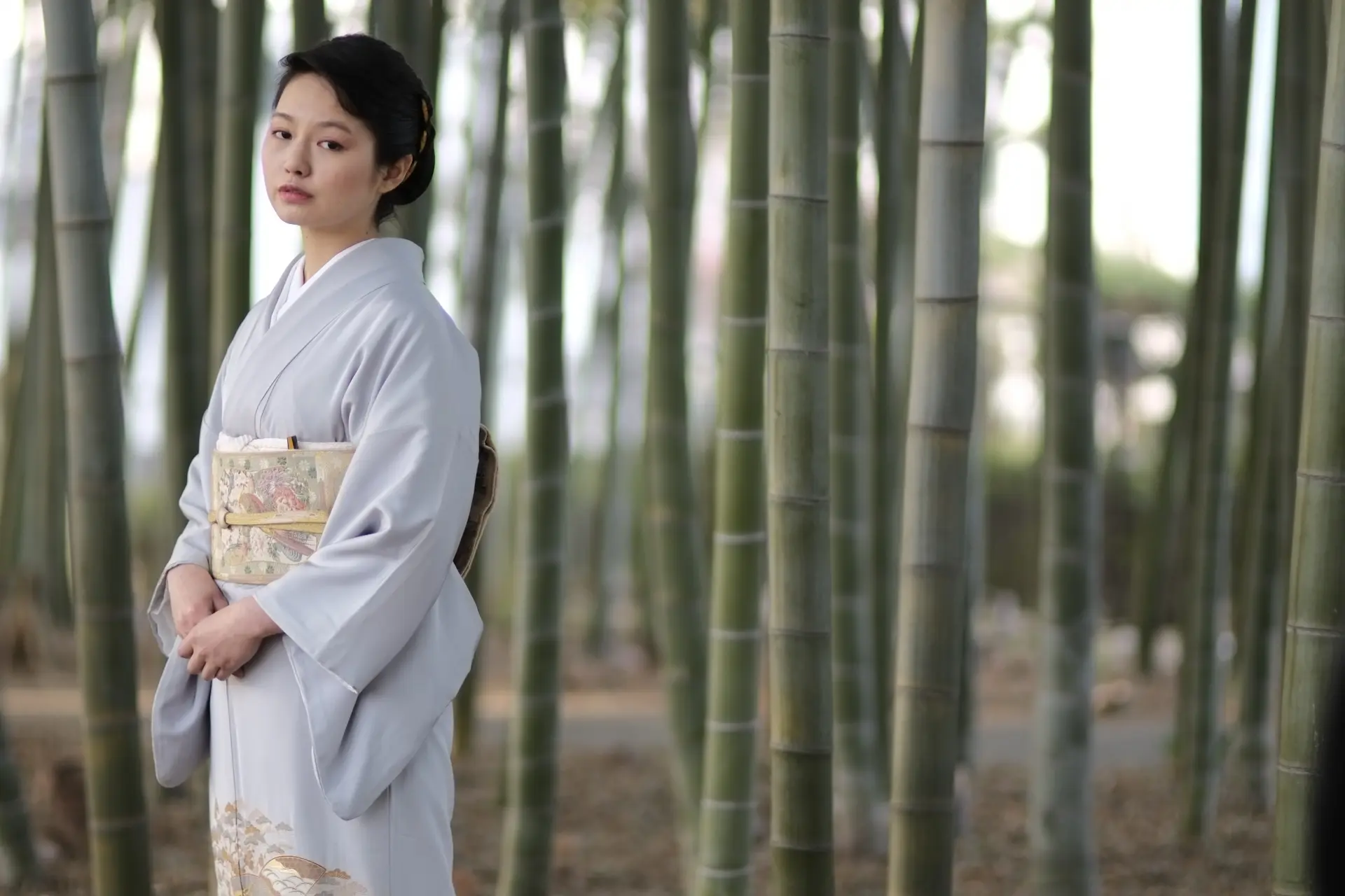 A young woman in a light gray kimono stands in a bamboo forest, looking forward.