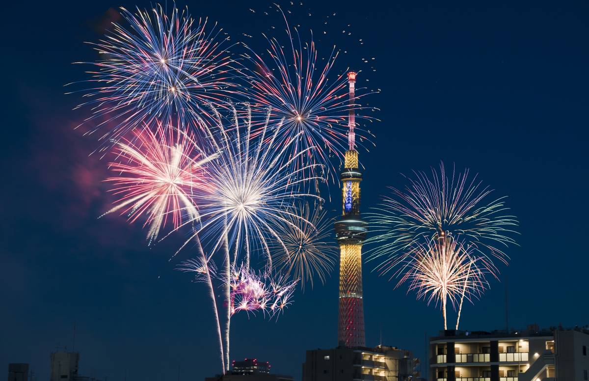Colorful fireworks burst around an illuminated tower and city buildings at night.