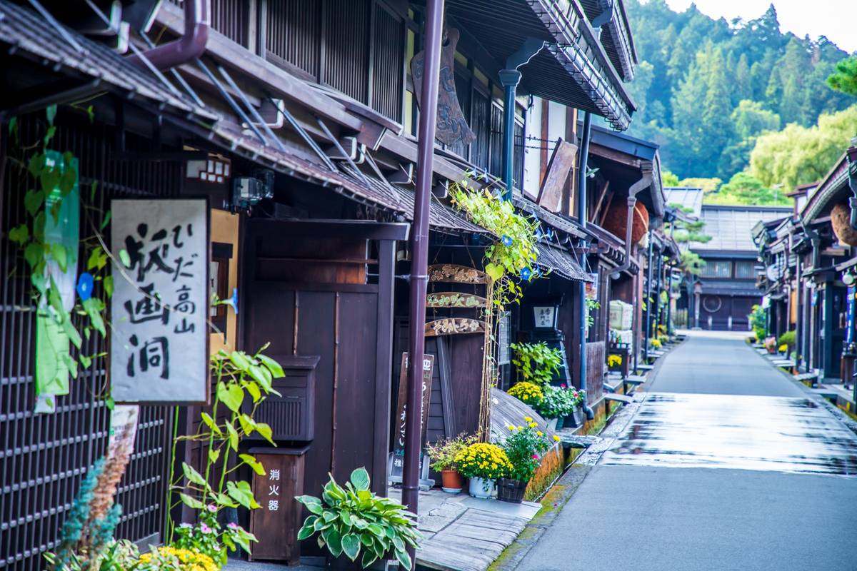 A narrow, wet street lined with traditional dark wooden Japanese buildings adorned with plants and flowers, with green hills