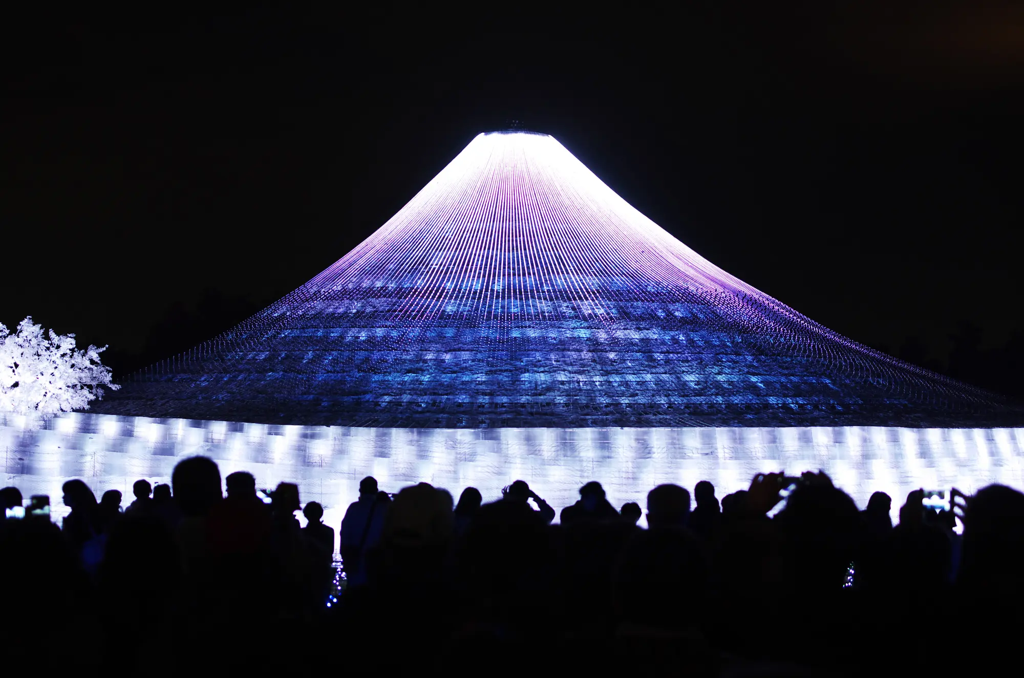 Conical light installation glowing purple and blue at night, with a silhouetted crowd watching.