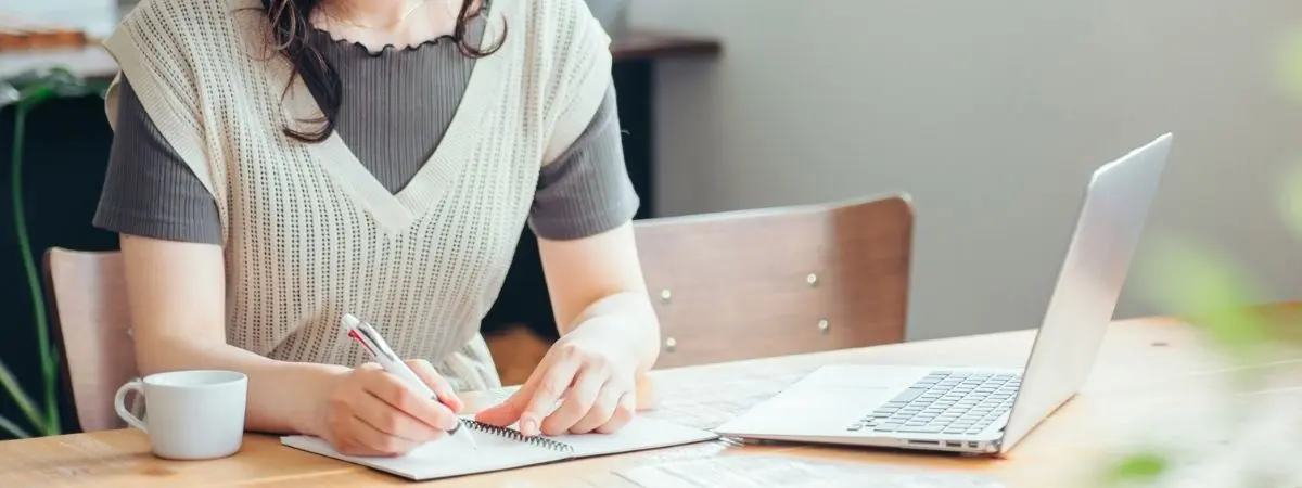A woman writing in a notebook with a laptop and mug on a wooden table.