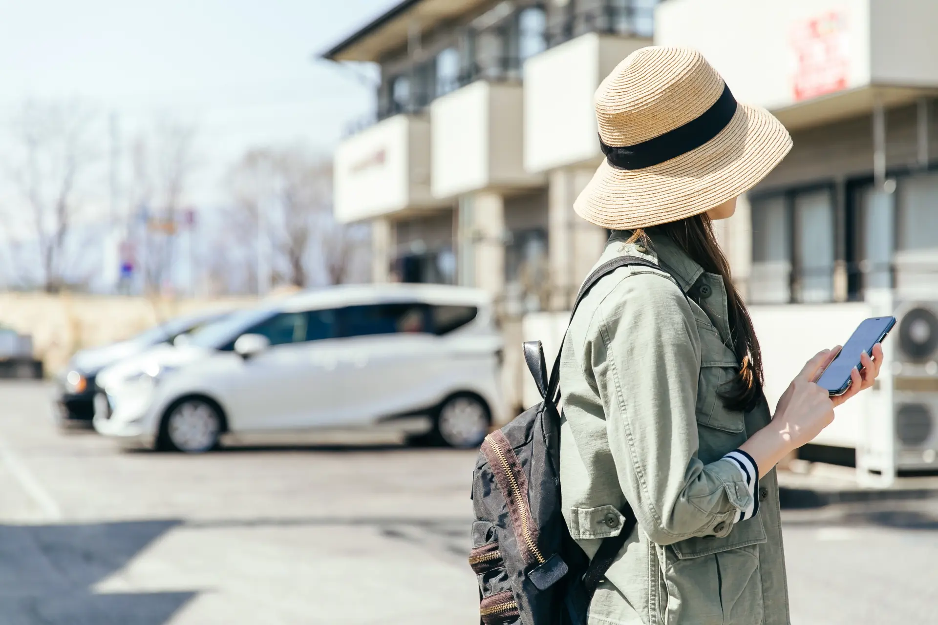 A woman in a straw hat and jacket looks at her smartphone outdoors.