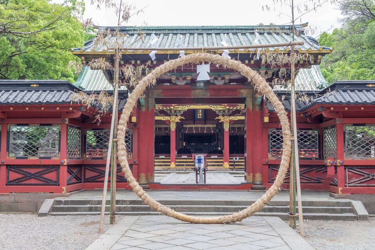 A large straw ring stands on a paved path leading to a traditional red and gold Japanese shrine.