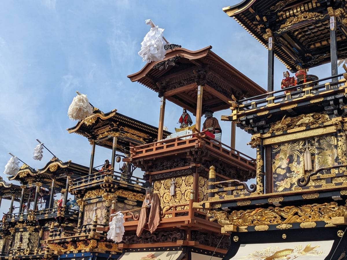 Several ornate black and gold Japanese festival floats with figures under a blue sky.