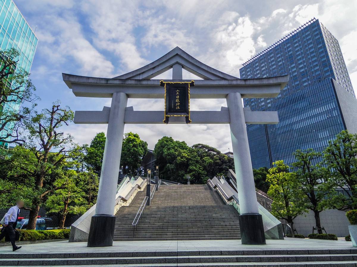 A large white torii gate with a black and gold plaque stands before a long stone staircase flanked by trees, with modern skyscrapers in the background.