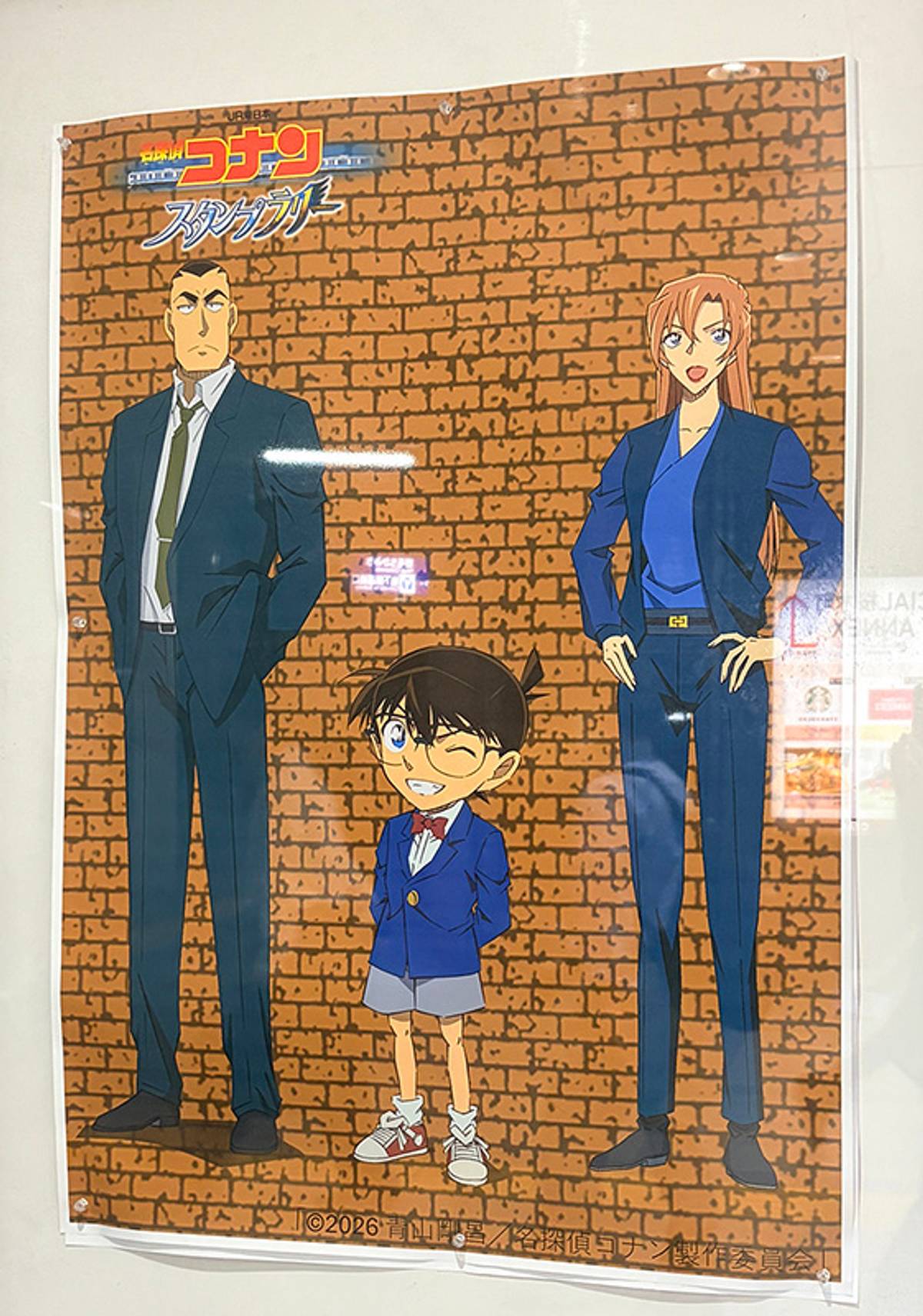 A poster for Detective Conan featuring a winking Conan Edogawa, a tall man in a suit, and a woman in a blue outfit, against a brick wall.