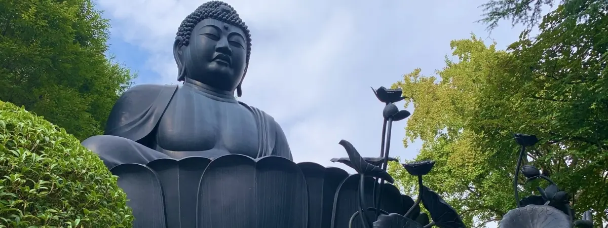 Large dark Buddha statue seated among green foliage and lotus sculptures.
