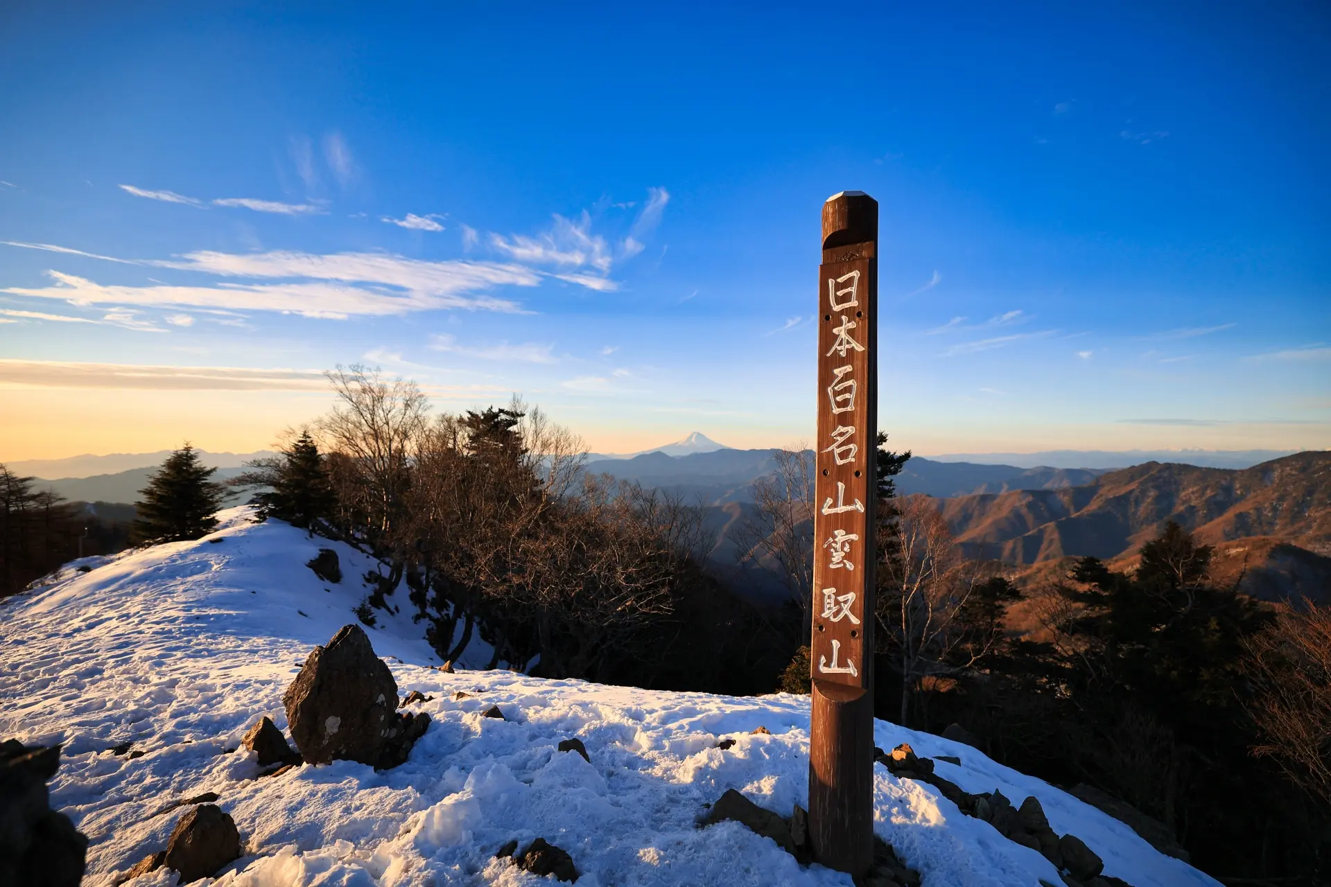Snow-covered mountain peak with a wooden signpost in Japanese, distant mountains, and Mt. Fuji under a clear sky.