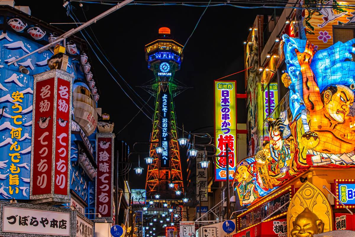 A vibrant night scene in Osaka with the illuminated Tsutenkaku Tower rising above streets lined with colorful neon signs and ornate glowing figures.