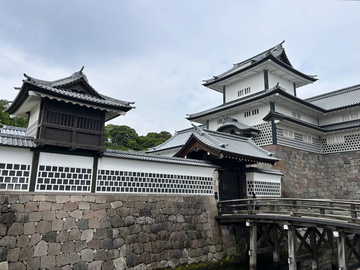 A Japanese castle complex with white walls, dark roofs, a stone foundation, and a wooden bridge over a moat.