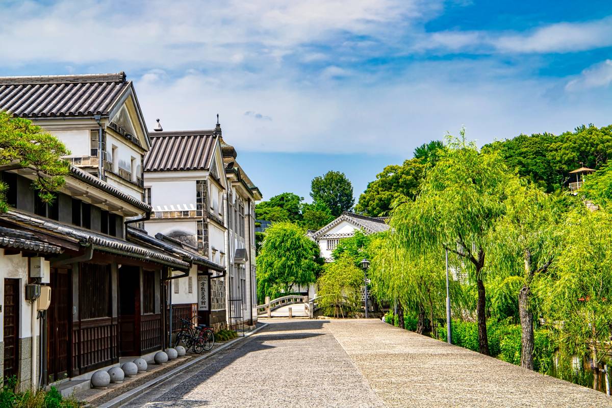 Traditional white Japanese buildings line a stone path next to a canal with weeping willow trees.