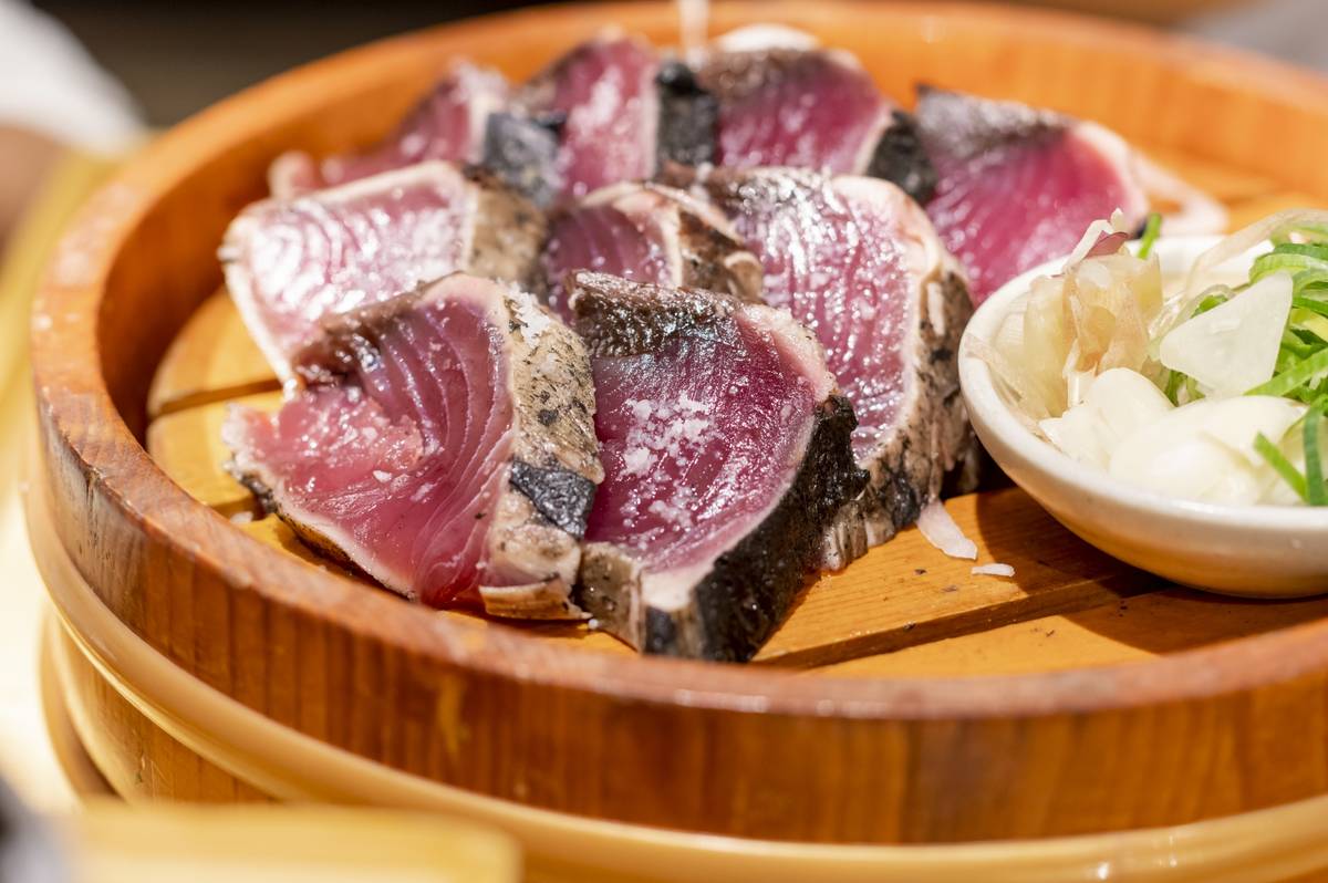 Slices of seared tuna in a wooden bowl with a side of green onions and other garnishes.