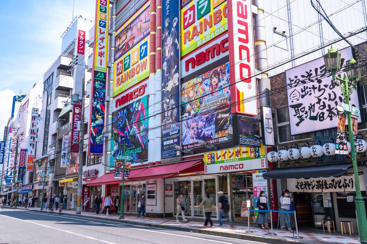 A vibrant Japanese city street lined with buildings covered in colorful arcade and anime advertisements, with people walking on the sidewalk.