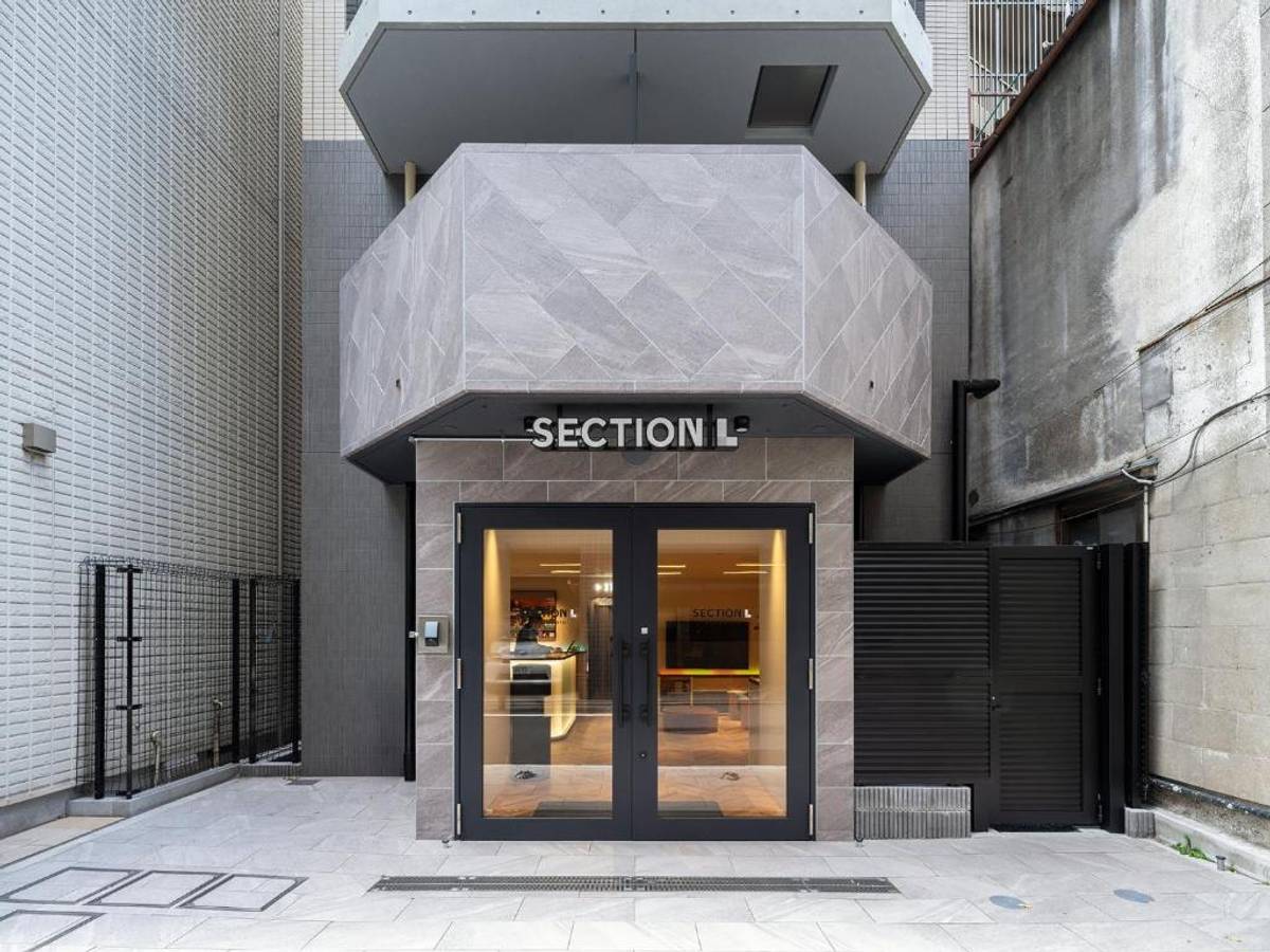 Modern entrance of a building named "SECTION L" with glass doors, bordered by a white tiled wall on the left and a rough concrete wall on the right.