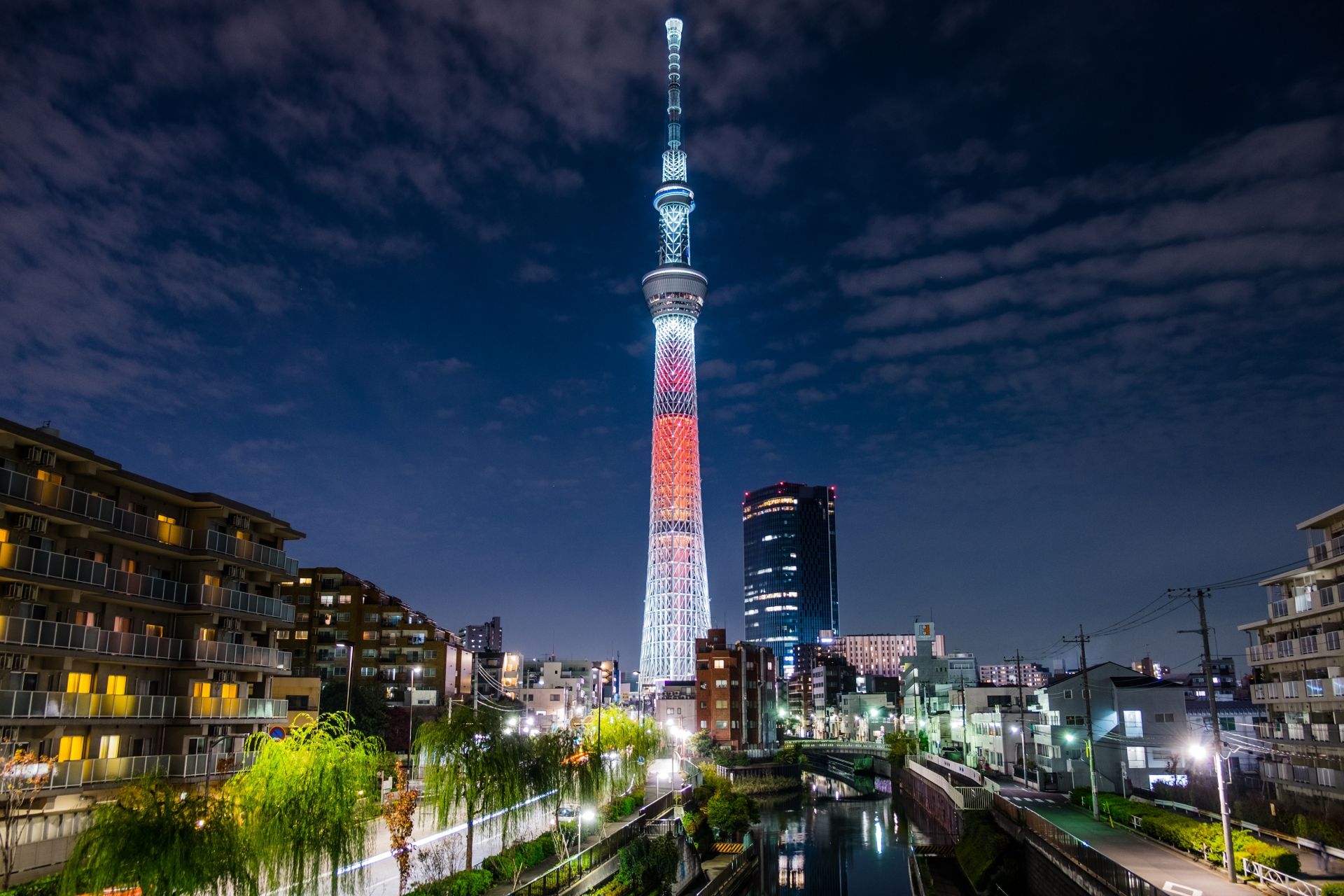 Tokyo Skytree illuminated red and white at night, overlooking a city with a canal.