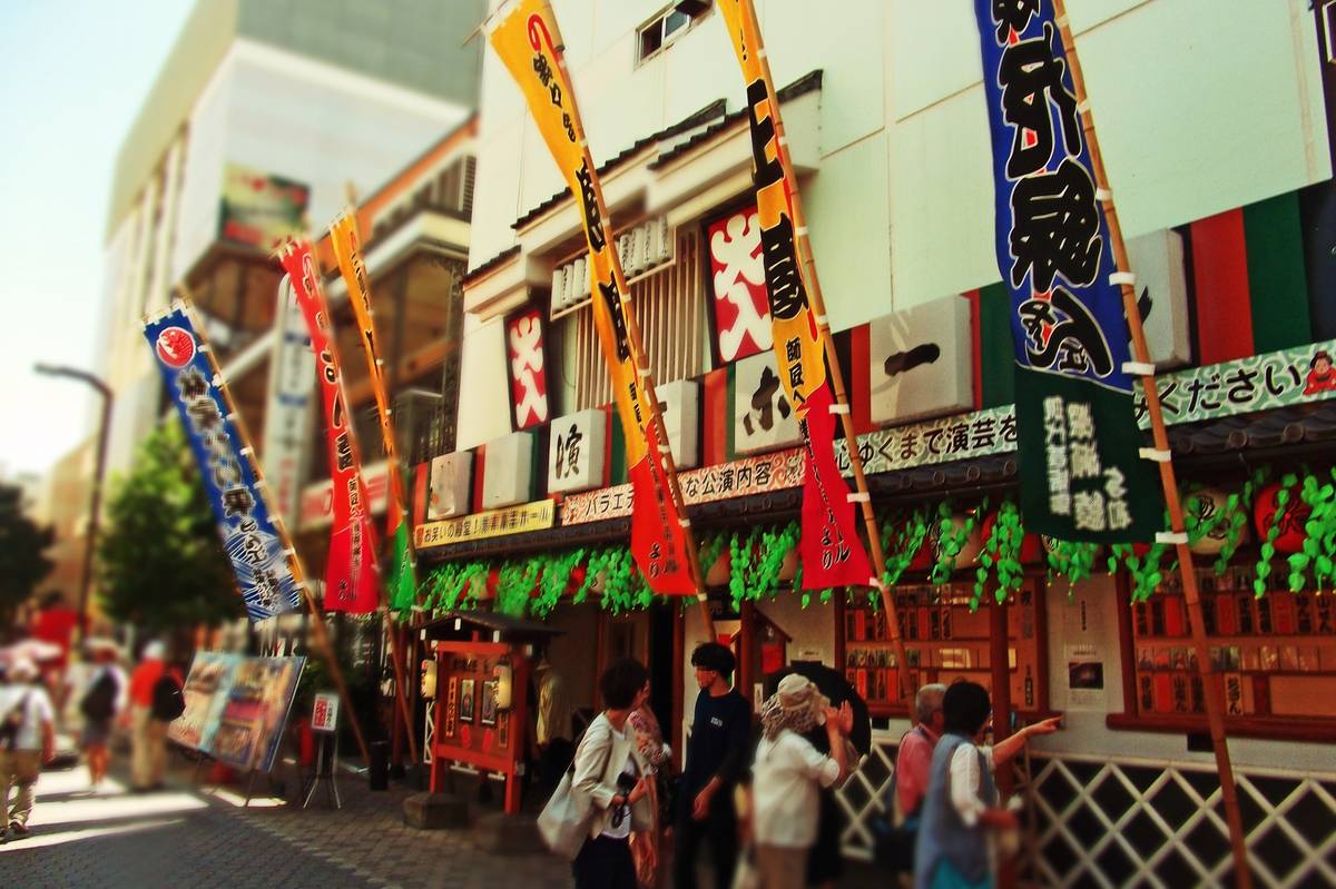 A Japanese street with shops decorated by many colorful banners and people walking, rendered with a tilt-shift effect.