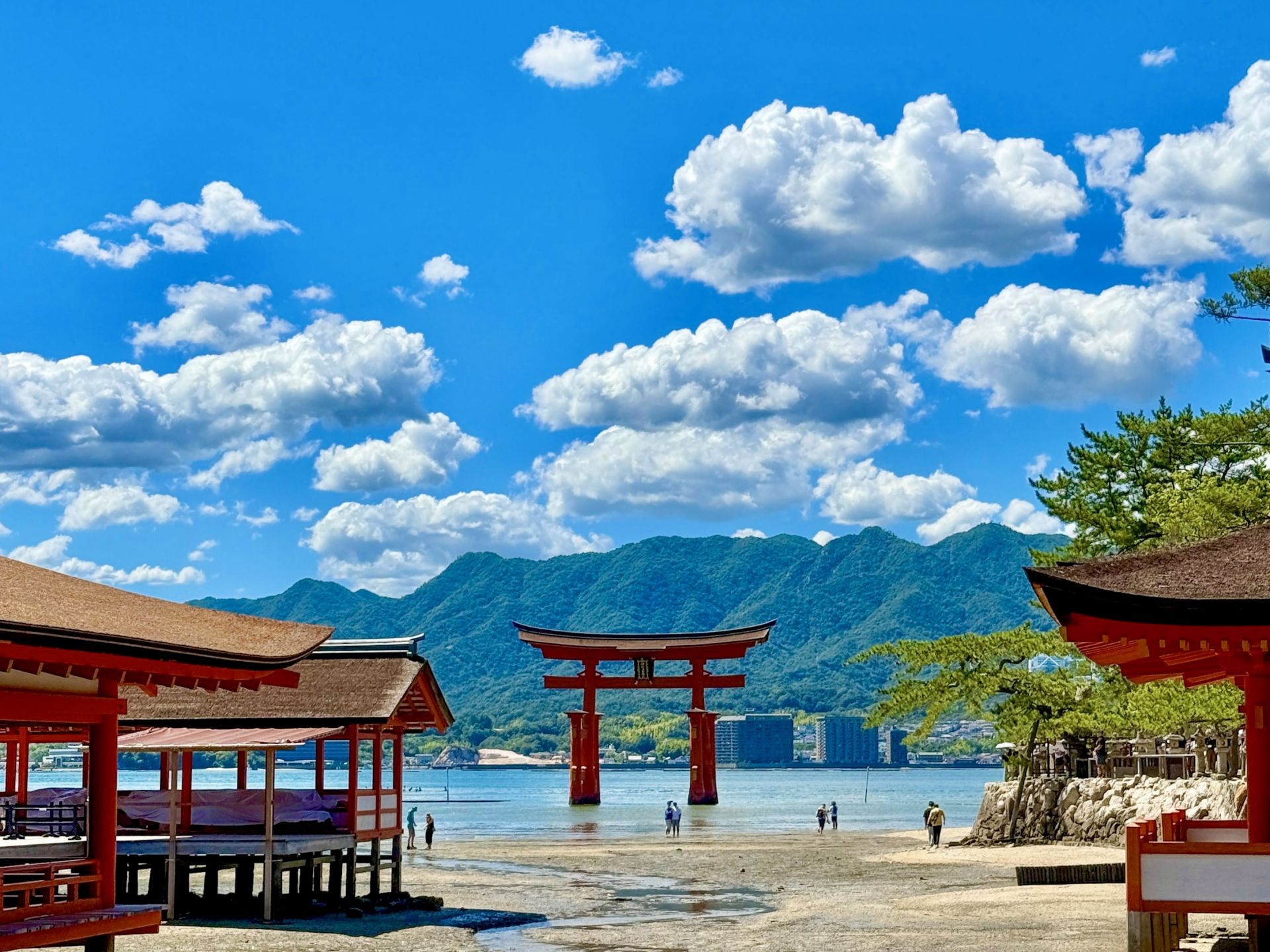 Red torii gate in water at Itsukushima Shrine, with shrine buildings on the shore, mountains, and a cloudy blue sky.
