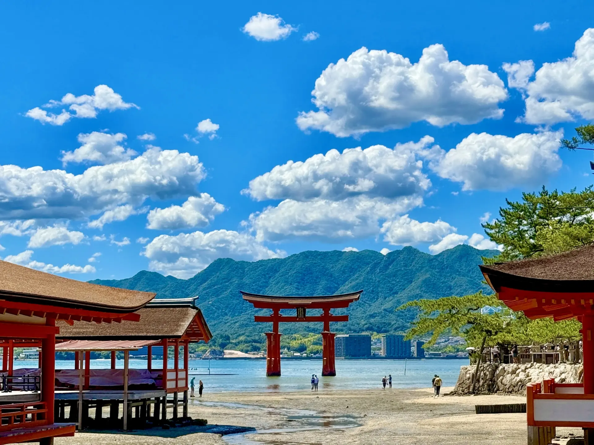 Red torii gate in water at Itsukushima Shrine, with shrine buildings on the shore, mountains, and a cloudy blue sky.