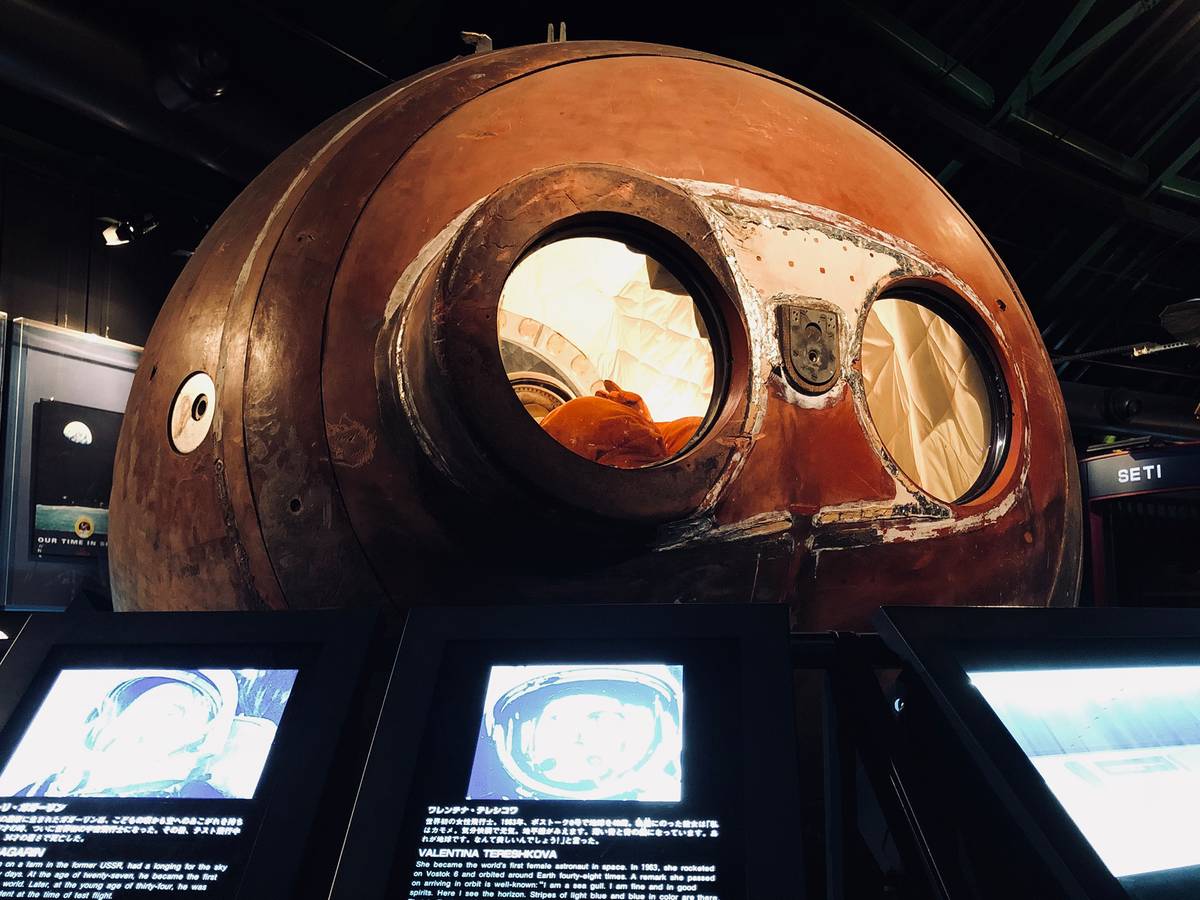 A large, weathered, reddish-brown space capsule with two circular windows sits above illuminated museum display screens.