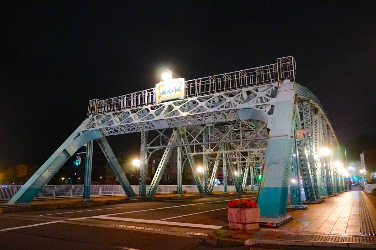 Illuminated light blue truss bridge at night, with a sign overhead.