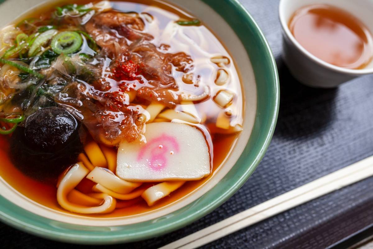 Close-up of a bowl of udon noodles topped with fish cake, scallions, and mushrooms, beside a cup of tea and chopsticks.