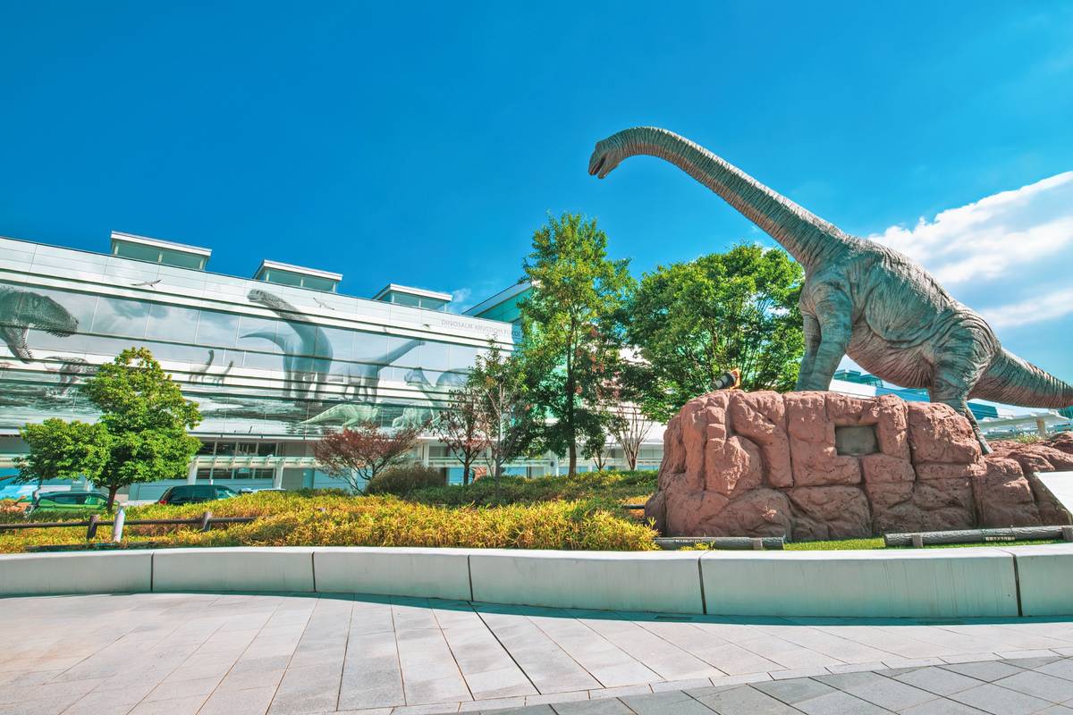A large long-necked dinosaur statue stands outside a modern building with dinosaur images on its windows, under a clear blue sky.