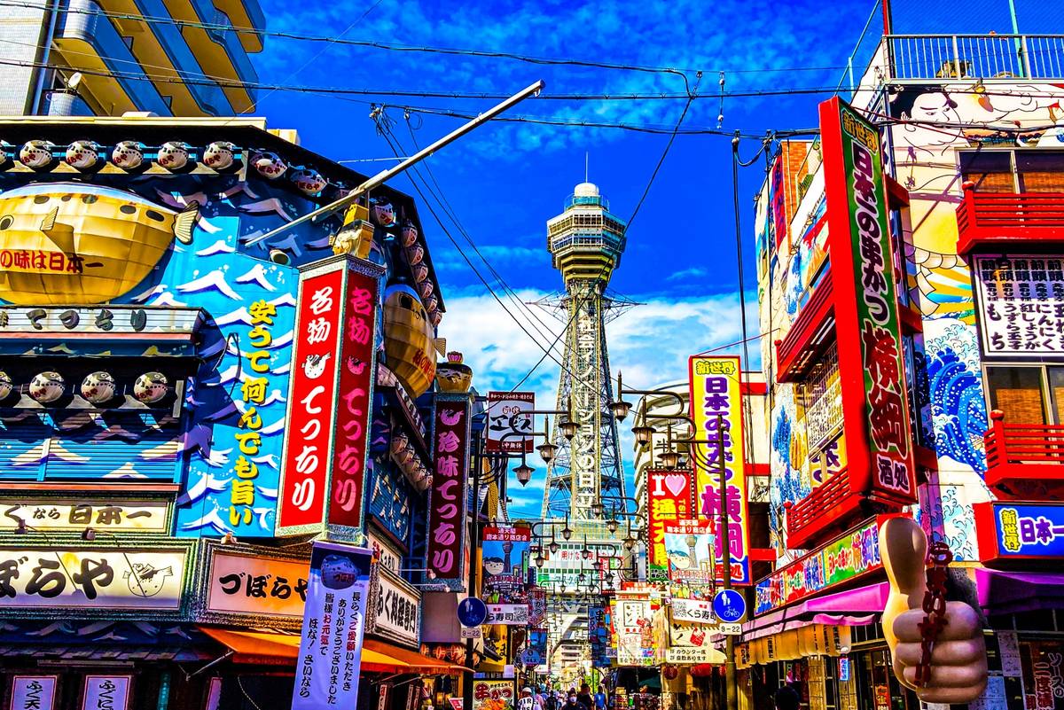 Vibrant street in Osaka, Japan, lined with colorful signs, with Tsutenkaku Tower in the background.