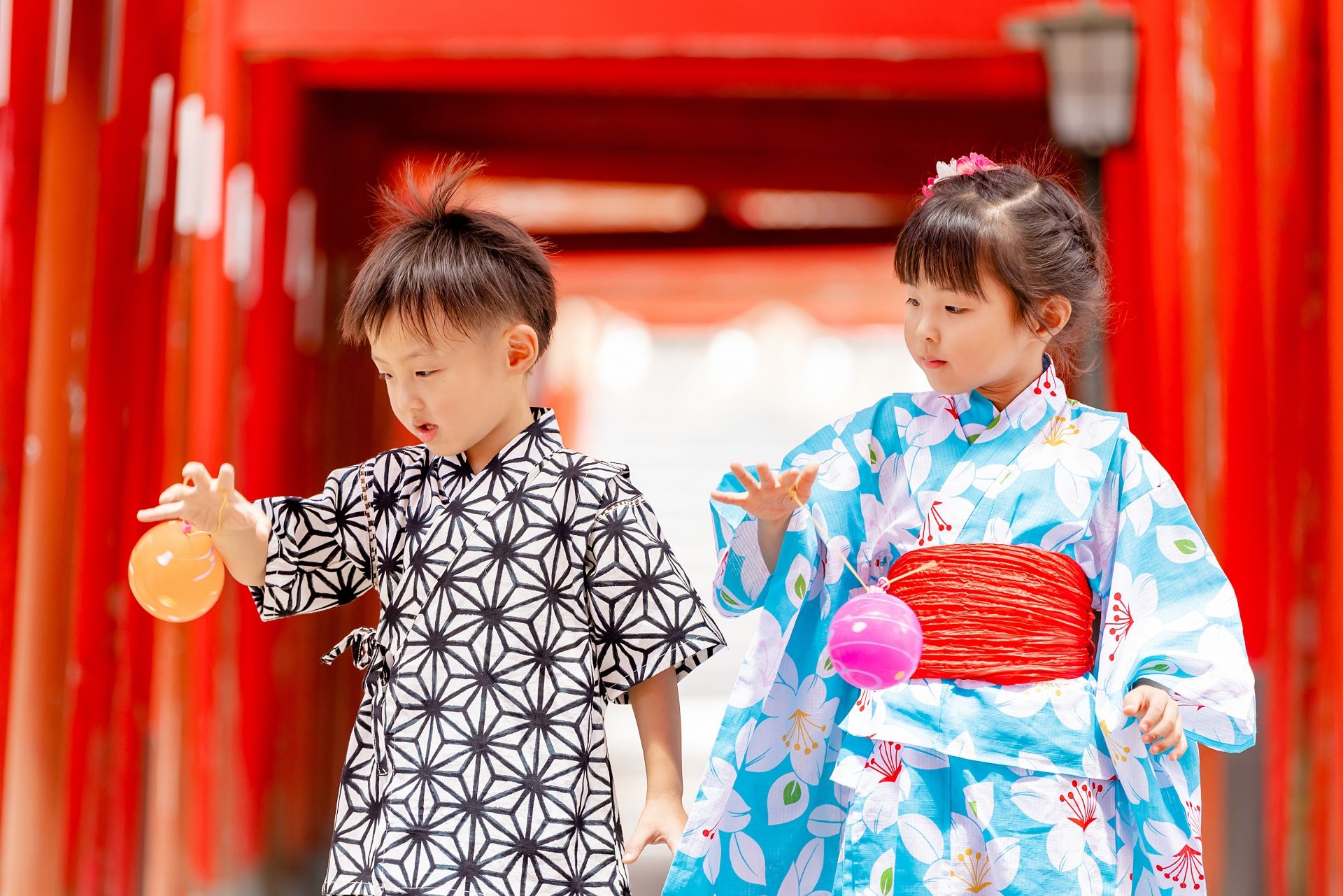 Two young children in yukata play with water balloons in front of red torii gates.