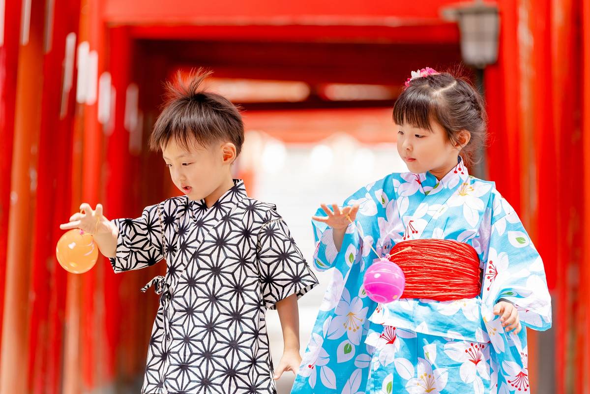 Two young children in yukata play with water balloons in front of red torii gates.