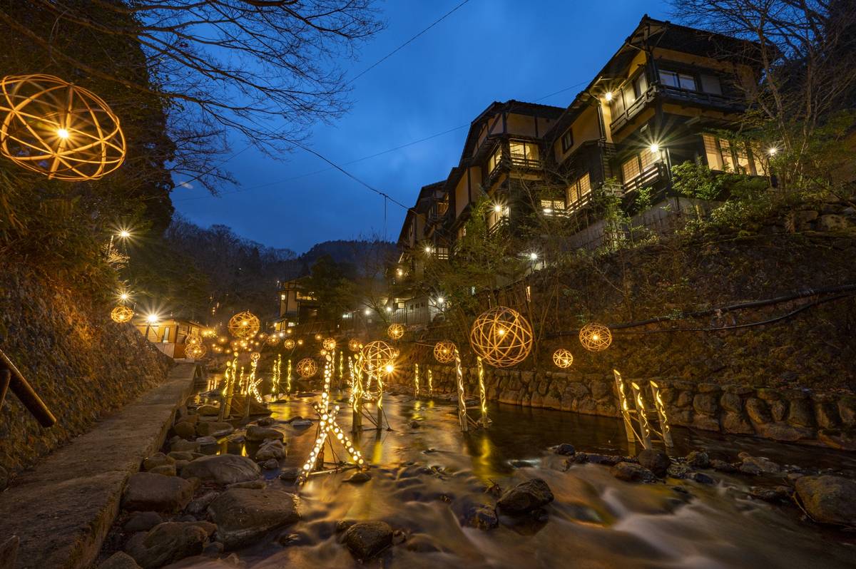 A scenic view at dusk shows a winding river illuminated by glowing spherical lanterns and lights on posts.