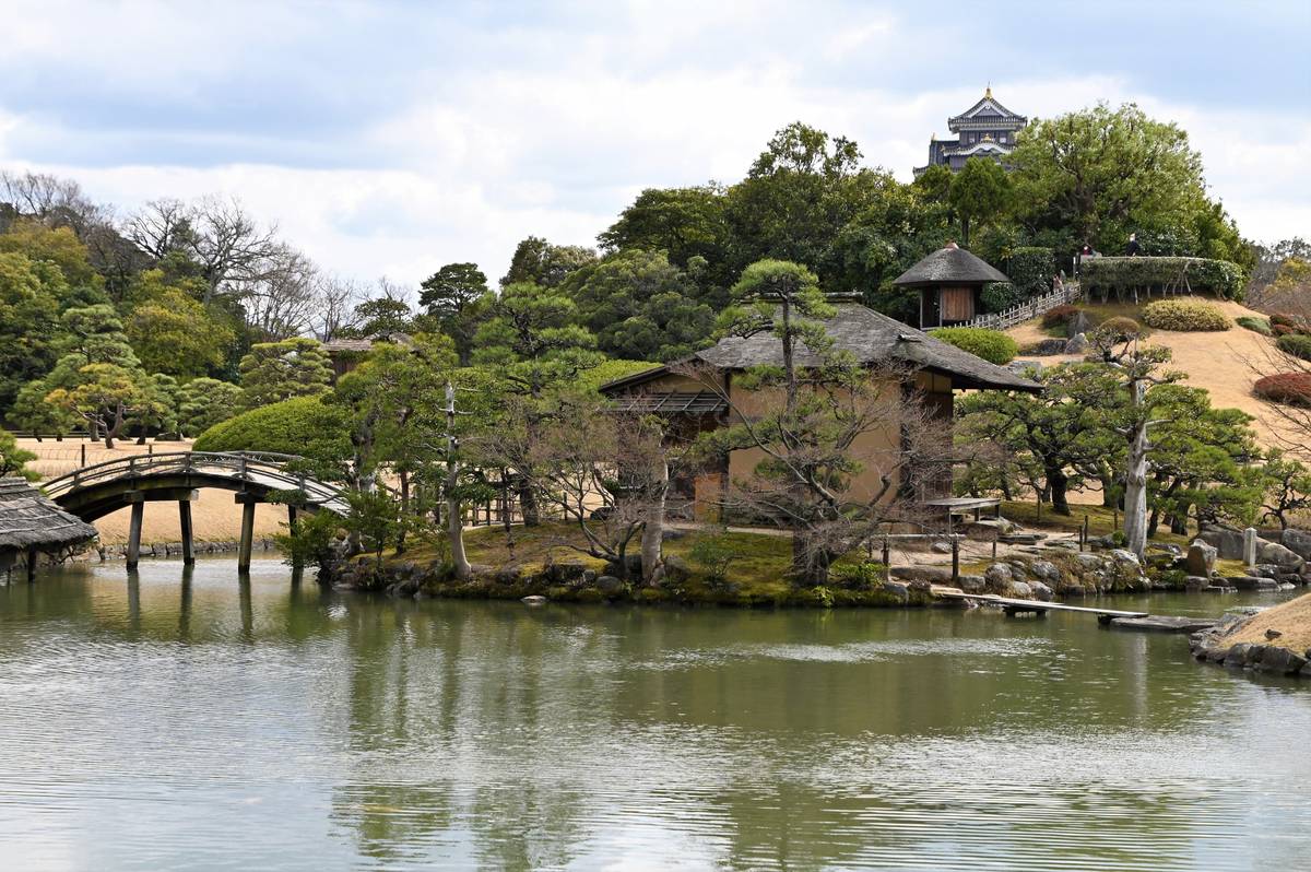 Japanese garden with a pond, arched bridge, traditional house, and a castle on a distant hill.
