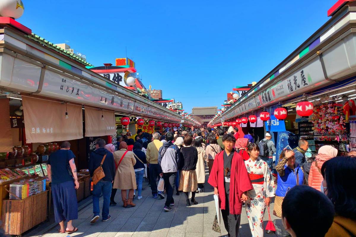 A lively, crowded pedestrian street lined with shops, some displaying red lanterns, with many people walking, including a couple in traditional Japanese kimonos.
